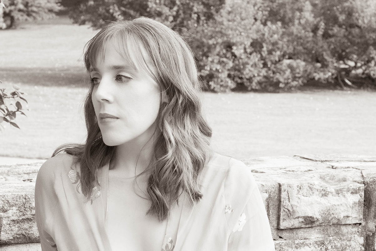 Black and white headshot of Leslee Wood leaning against a stone wall, looking thoughtfully toward the side.